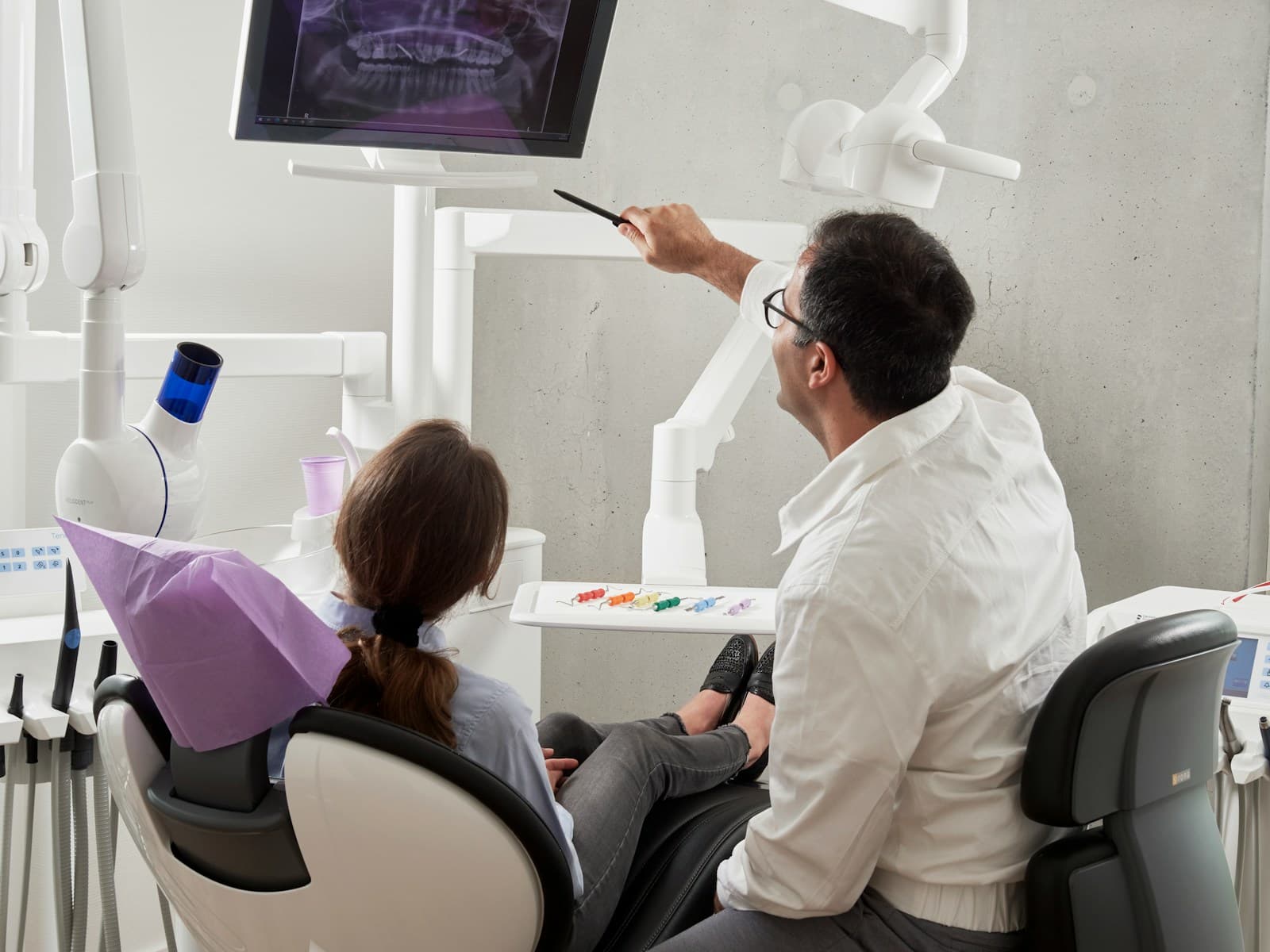 A dentist pointing at an X-ray on a screen, explaining it to a patient in the chair