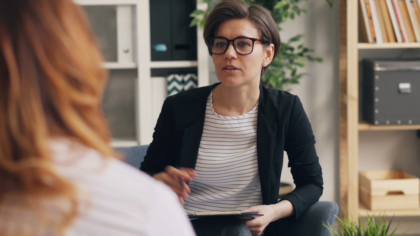 A counsellor with a notepad listening to a client in a calm room with shelves and plants