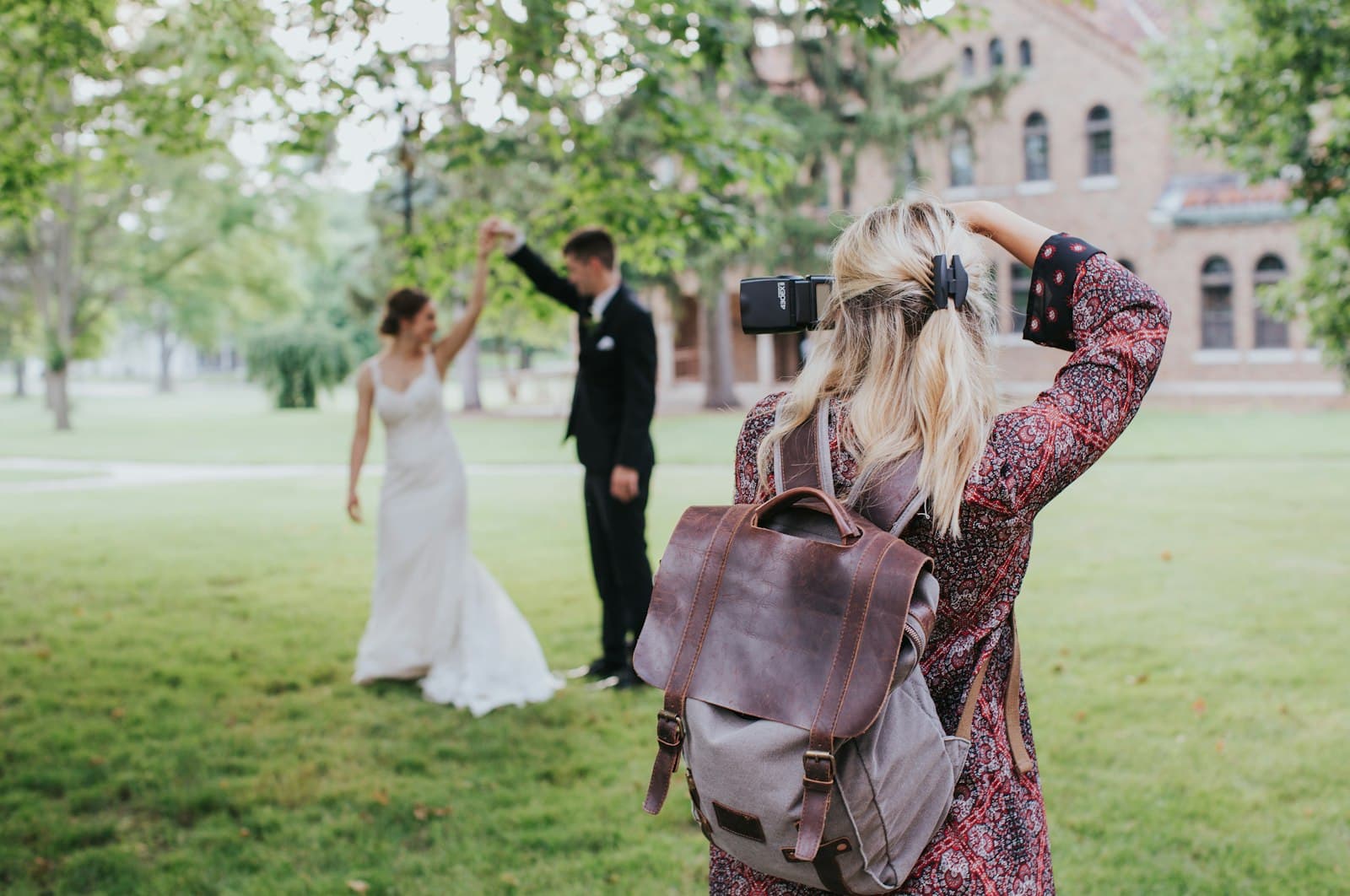 A wedding photographer with a camera and a leather backpack photographing a bride and groom dancing on a lawn